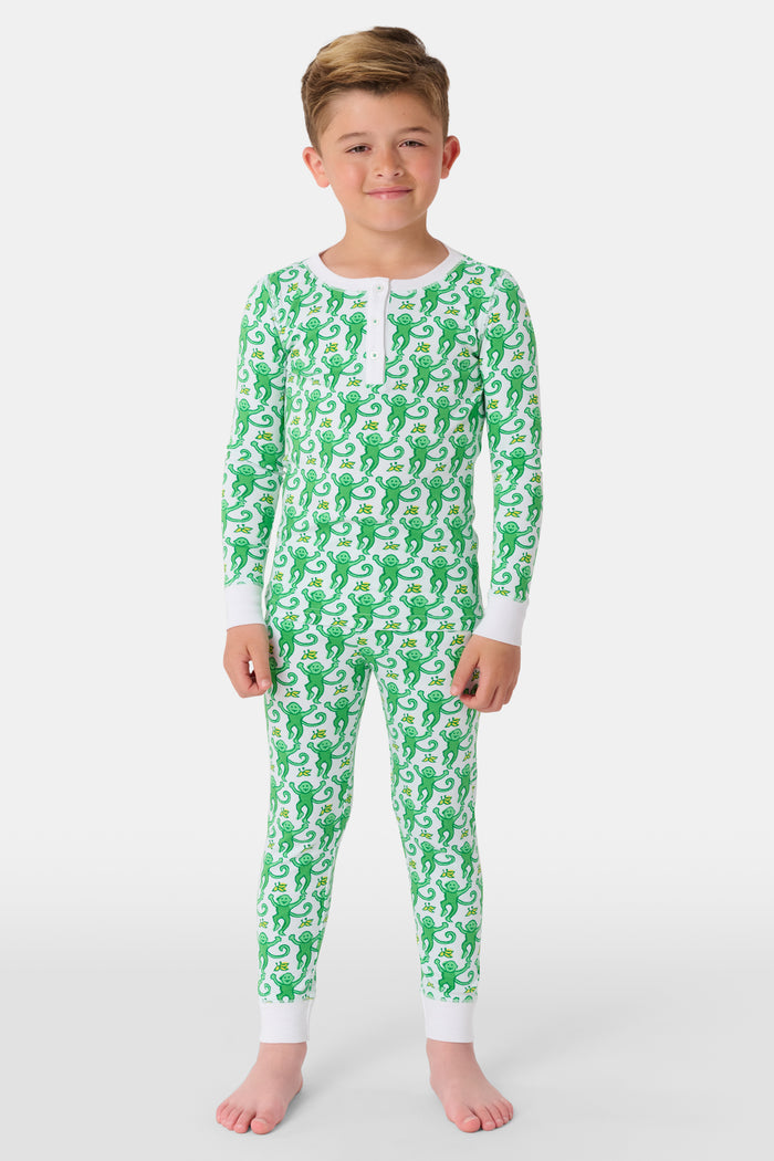 A young boy with light brown hair smiles barefoot in Roller Rabbit’s Kids Monkey Pajamas—a matching 100 cotton set with playful monkey print, white cuffs, and a buttoned neckline—against a plain white background.