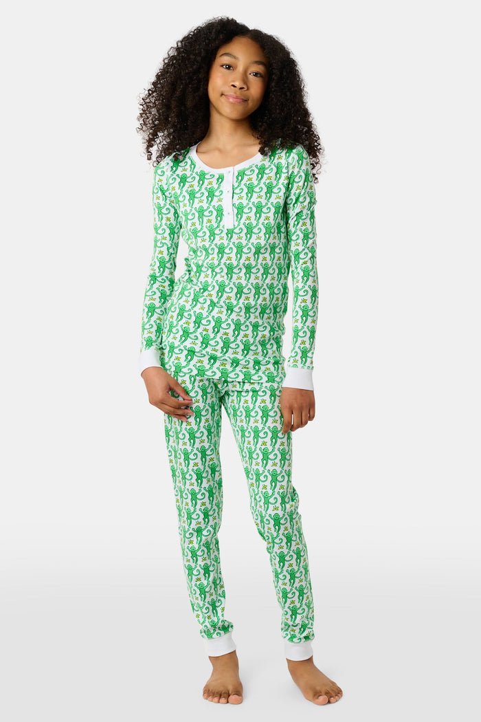 A young boy with light brown hair smiles barefoot in Roller Rabbit’s Kids Monkey Pajamas—a matching 100 cotton set with playful monkey print, white cuffs, and a buttoned neckline—against a plain white background.