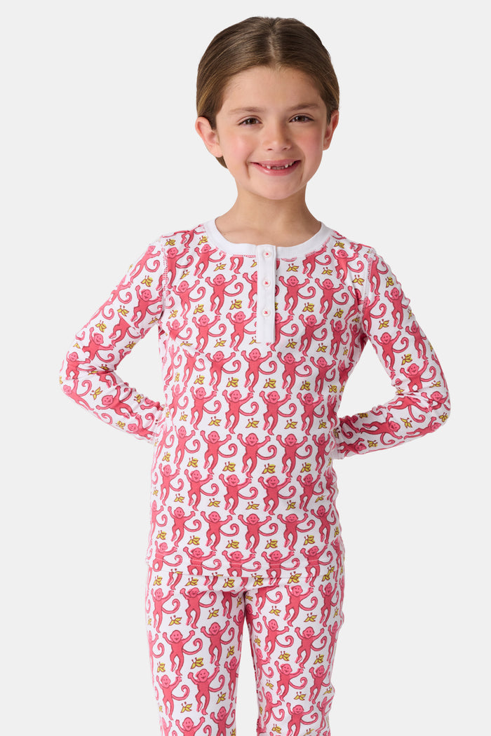 A young child smiles in Roller Rabbit Kids Monkey Pajamas—a matching long-sleeve, full-length set made from 100 Pima cotton with a playful monkey pattern and white cuffs, set against a plain white background.