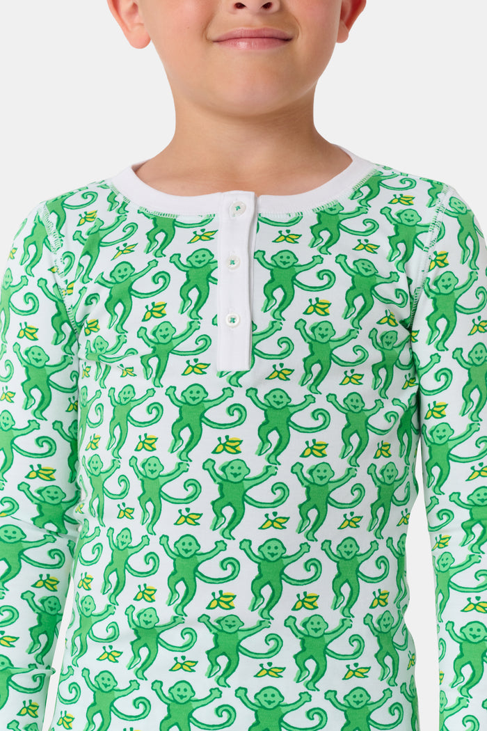 A young boy with light brown hair smiles barefoot in Roller Rabbit’s Kids Monkey Pajamas—a matching 100 cotton set with playful monkey print, white cuffs, and a buttoned neckline—against a plain white background.