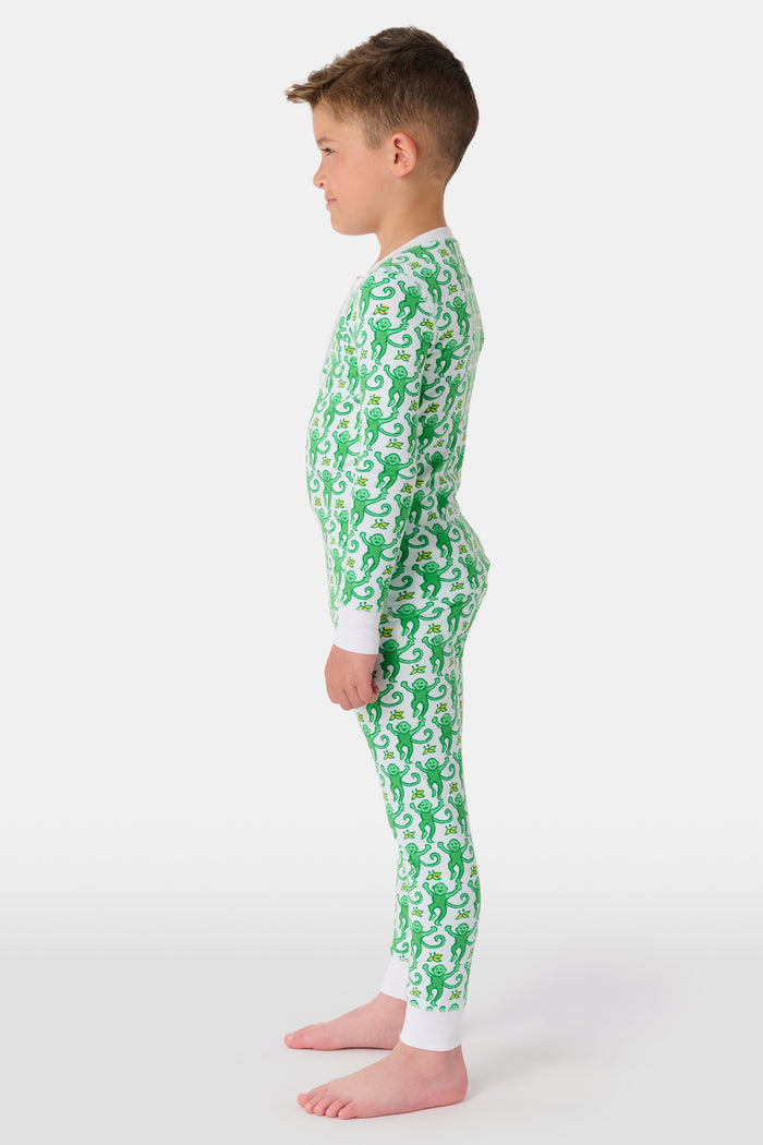 A young boy with light brown hair smiles barefoot in Roller Rabbit’s Kids Monkey Pajamas—a matching 100 cotton set with playful monkey print, white cuffs, and a buttoned neckline—against a plain white background.