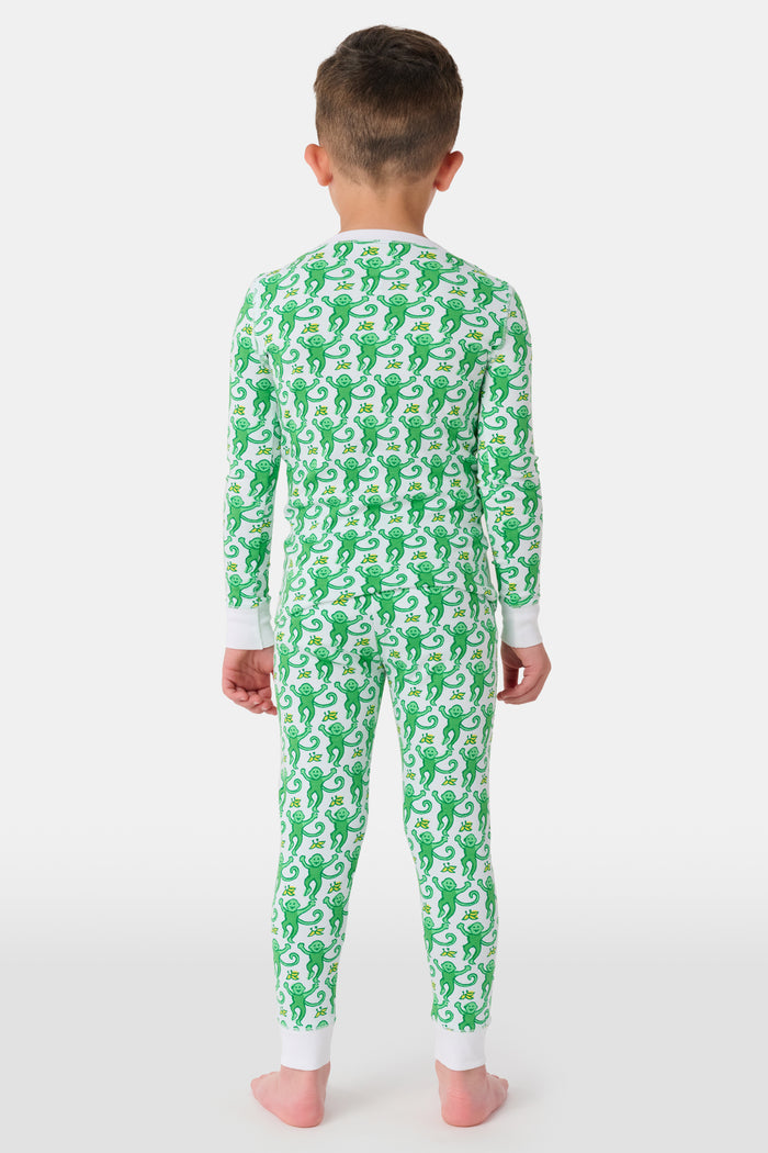 A young boy with light brown hair smiles barefoot in Roller Rabbit’s Kids Monkey Pajamas—a matching 100 cotton set with playful monkey print, white cuffs, and a buttoned neckline—against a plain white background.