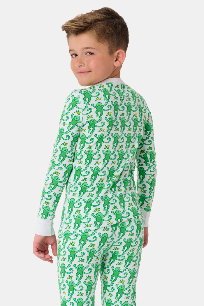 A young boy with light brown hair smiles barefoot in Roller Rabbit’s Kids Monkey Pajamas—a matching 100 cotton set with playful monkey print, white cuffs, and a buttoned neckline—against a plain white background.