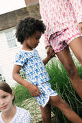 A child in the Roller Rabbit Kids Monkey Playa Play Set walks with an adult in pink and white patterned clothing. In the foreground, another child with brown hair wears a white polka dot shirt.