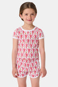 A young girl with brown hair wears the Roller Rabbit Kids Monkey Playa Play Set—pink and white pajamas featuring playful pink monkeys and yellow bananas—while smiling against a plain white background.