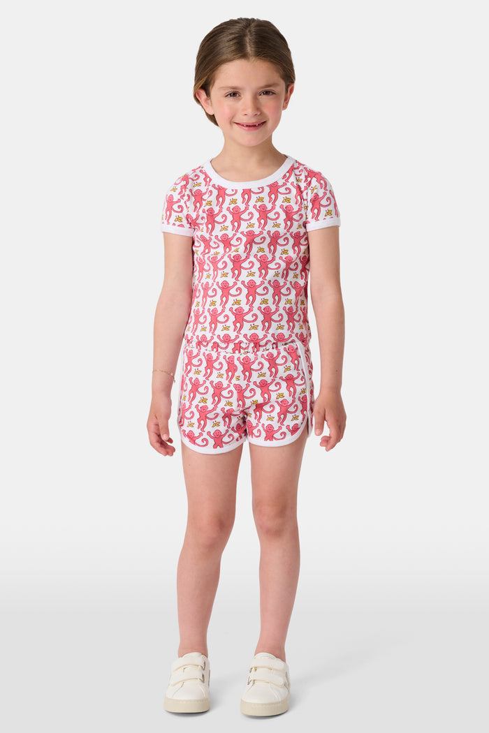 A young girl with brown hair wears the Roller Rabbit Kids Monkey Playa Play Set—pink and white pajamas featuring playful pink monkeys and yellow bananas—while smiling against a plain white background.