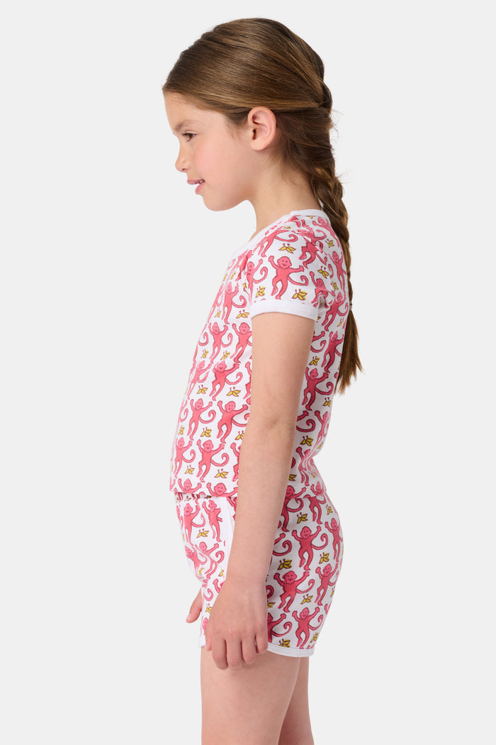 A young girl with brown hair wears the Roller Rabbit Kids Monkey Playa Play Set—pink and white pajamas featuring playful pink monkeys and yellow bananas—while smiling against a plain white background.