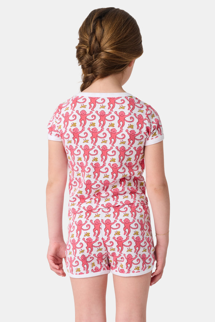 A young girl with brown hair wears the Roller Rabbit Kids Monkey Playa Play Set—pink and white pajamas featuring playful pink monkeys and yellow bananas—while smiling against a plain white background.
