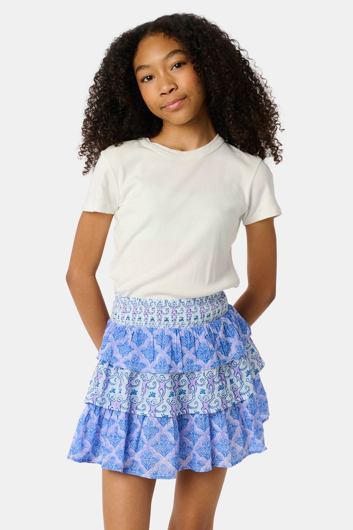 A young girl with curly hair wears a white t-shirt and the Roller Rabbit Kids Monkey/Jemina Sami Skirt, featuring ruffles and a smocked waist, standing against a light background with her hands behind her back and a slight smile.