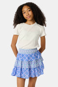 A young girl with curly hair wears a white t-shirt and the Roller Rabbit Kids Monkey/Jemina Sami Skirt, featuring ruffles and a smocked waist, standing against a light background with her hands behind her back and a slight smile.