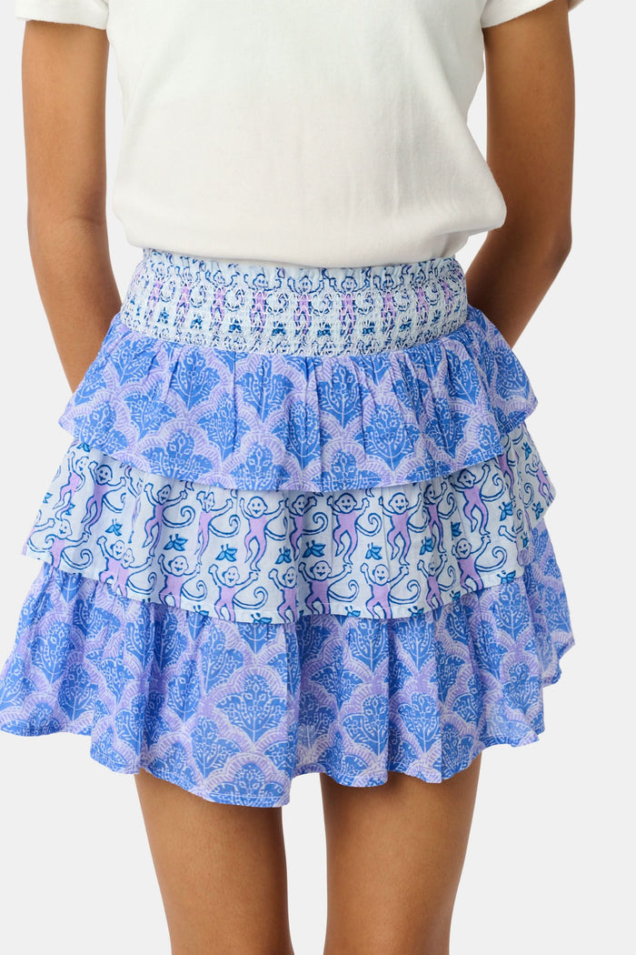 A young girl with curly hair wears a white t-shirt and the Roller Rabbit Kids Monkey/Jemina Sami Skirt, featuring ruffles and a smocked waist, standing against a light background with her hands behind her back and a slight smile.