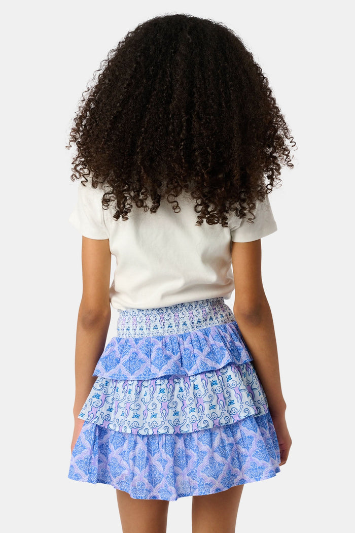 A young girl with curly hair wears a white t-shirt and the Roller Rabbit Kids Monkey/Jemina Sami Skirt, featuring ruffles and a smocked waist, standing against a light background with her hands behind her back and a slight smile.