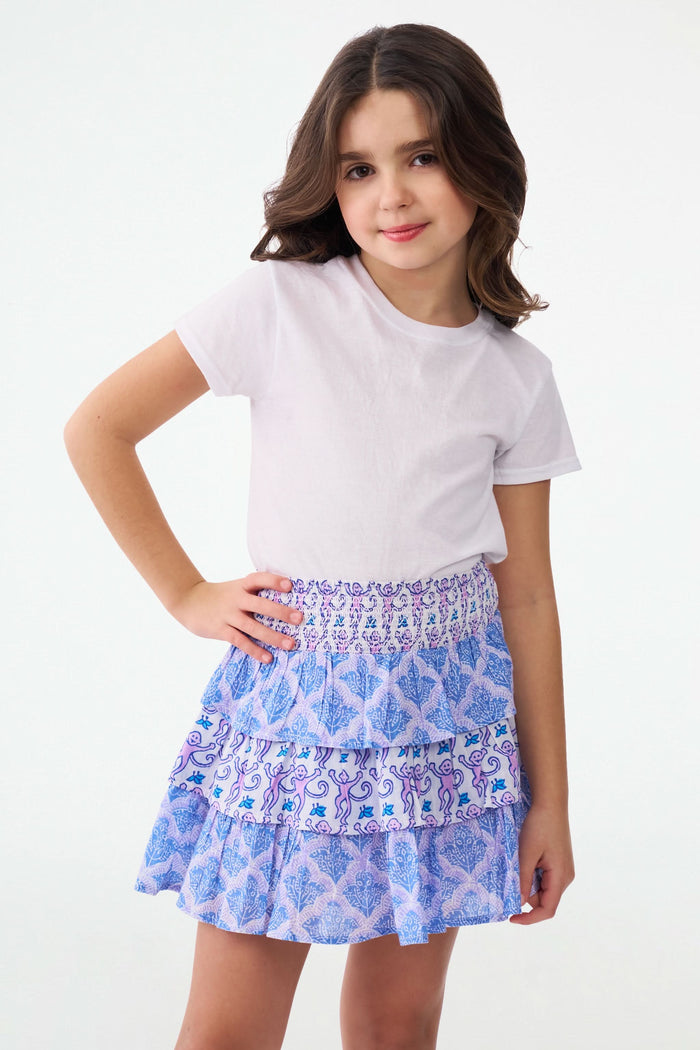 A young girl with curly hair wears a white t-shirt and the Roller Rabbit Kids Monkey/Jemina Sami Skirt, featuring ruffles and a smocked waist, standing against a light background with her hands behind her back and a slight smile.