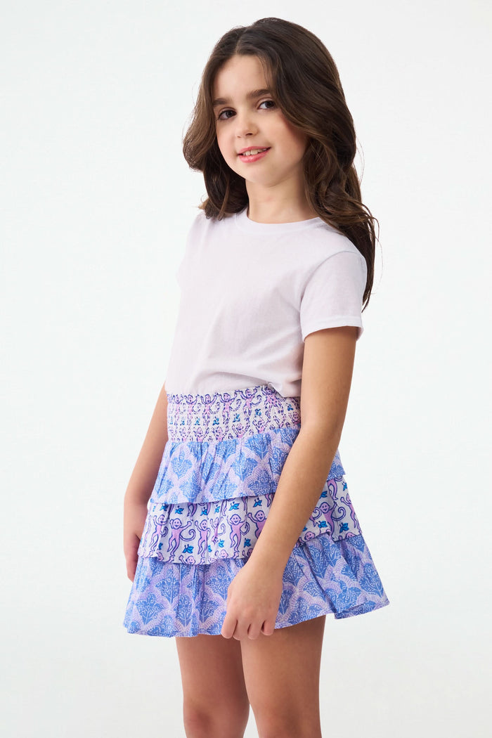 A young girl with curly hair wears a white t-shirt and the Roller Rabbit Kids Monkey/Jemina Sami Skirt, featuring ruffles and a smocked waist, standing against a light background with her hands behind her back and a slight smile.