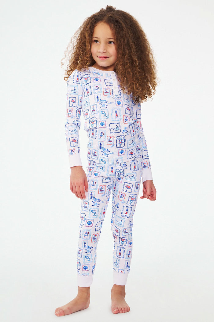 A young girl with curly hair stands barefoot, wearing Roller Rabbit Kids Nantucket Memories Pajamas crafted from soft Pima cotton, featuring a colorful whimsical print, set against a simple white background.
