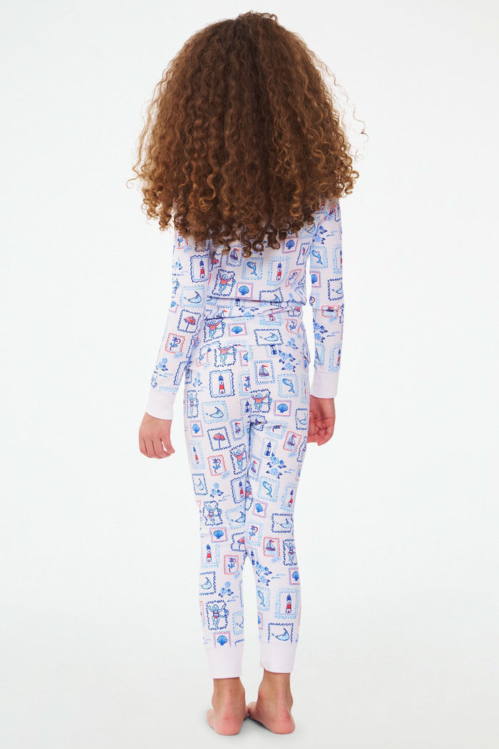 A young girl with curly hair stands barefoot, wearing Roller Rabbit Kids Nantucket Memories Pajamas crafted from soft Pima cotton, featuring a colorful whimsical print, set against a simple white background.