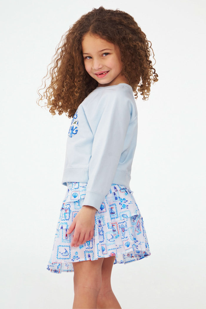 A child in a light blue sweatshirt and the Roller Rabbit Kids Nantucket Memories Sami Skirt, featuring blue and pink nautical patterns, stands with one hand on the tiered ruffle skirt against a plain white background.
