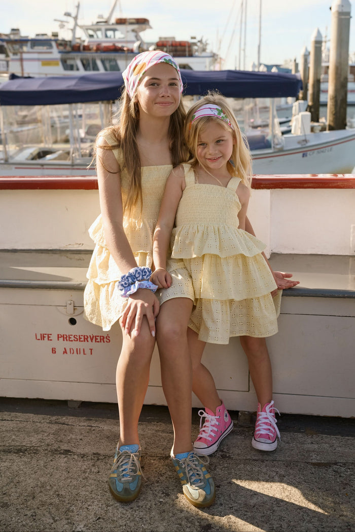 Two young girls smile at the camera on a dock in front of boats, both wearing matching Roller Rabbit Kids Petal Eyelet Kenia Dresses. The older girl sports sneakers and a blue scrunchie, while the younger wears pink shoes.