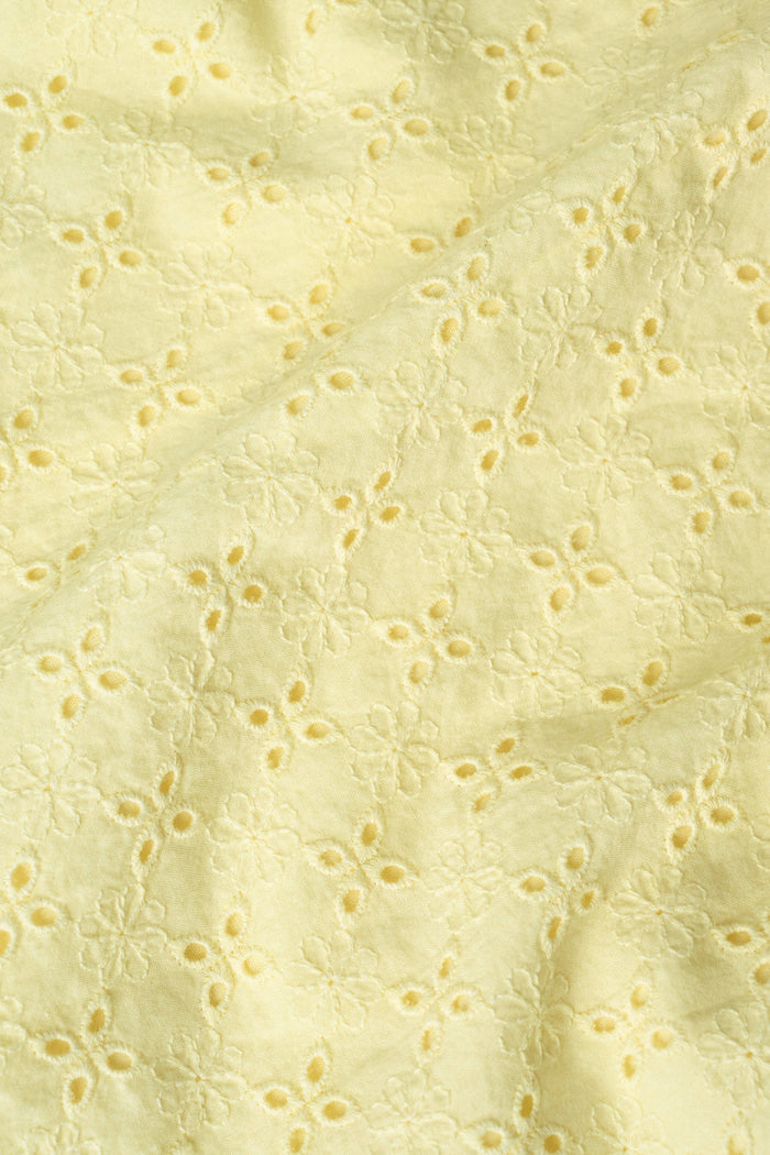 A woman with curly hair wears the Roller Rabbit Petal Eyelet Aurelia Top, a yellow cotton camisole with eyelet details and a ruffled hem, paired with a matching high-waisted skirt against a plain light background.