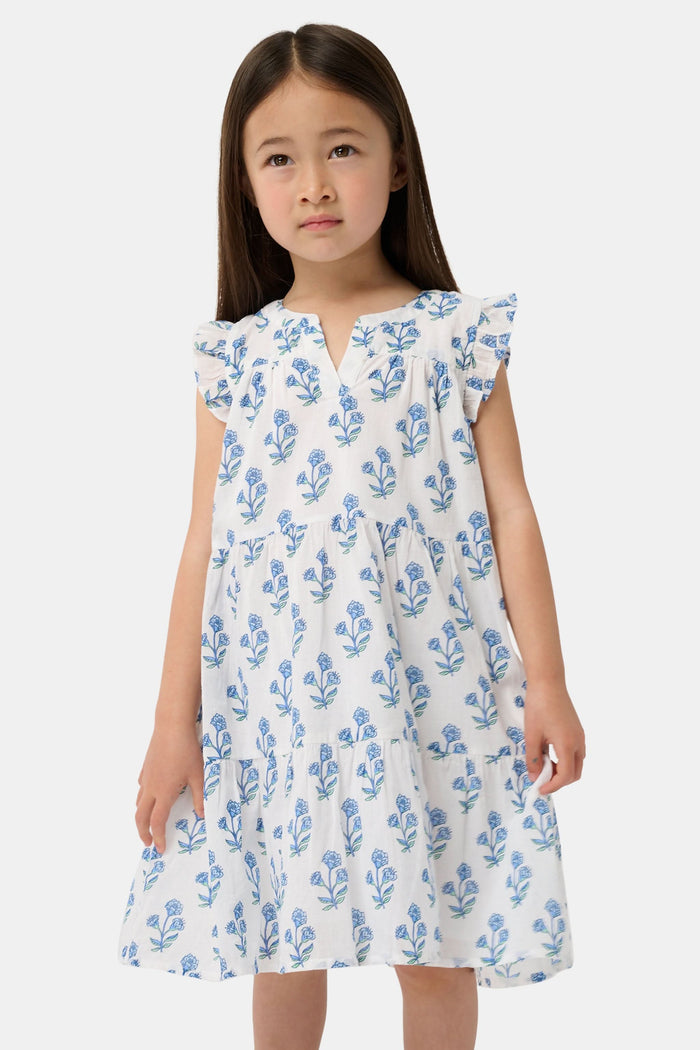 A young girl with long brown hair wears the Roller Rabbit Kids Rosa Ditsy Floral Pami Dress, made from cotton voile with blue-green block print and ruffled sleeves, standing against a plain light background, looking slightly to the side.