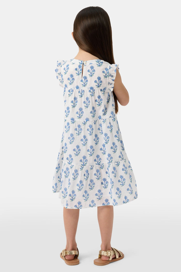 A young girl with long brown hair wears the Roller Rabbit Kids Rosa Ditsy Floral Pami Dress, made from cotton voile with blue-green block print and ruffled sleeves, standing against a plain light background, looking slightly to the side.