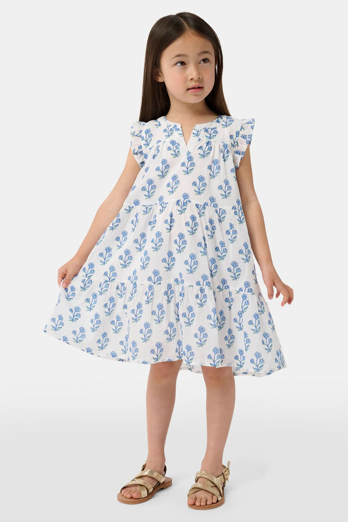 A young girl with long brown hair wears the Roller Rabbit Kids Rosa Ditsy Floral Pami Dress, made from cotton voile with blue-green block print and ruffled sleeves, standing against a plain light background, looking slightly to the side.