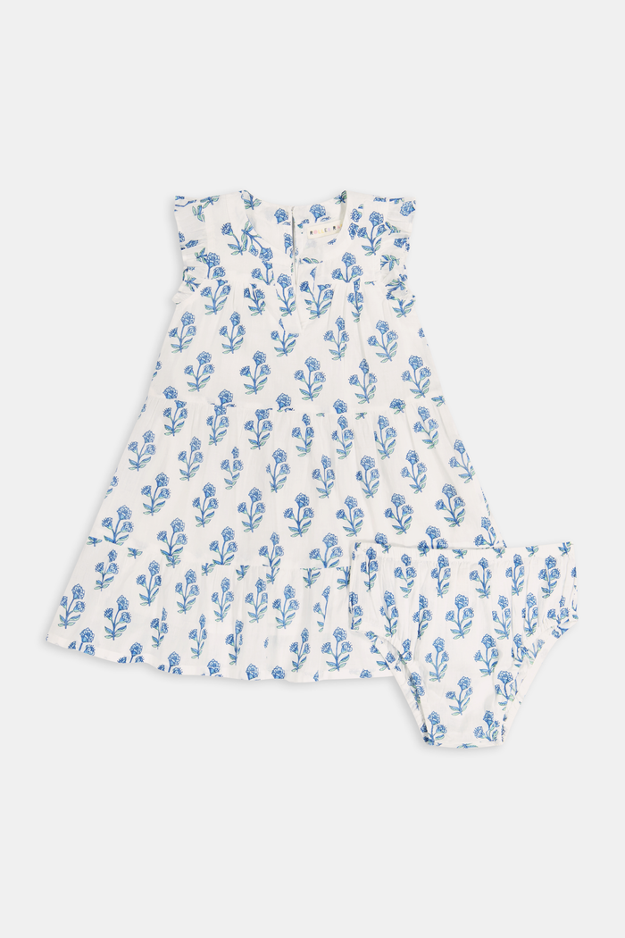 A young girl with long brown hair wears the Roller Rabbit Kids Rosa Ditsy Floral Pami Dress, made from cotton voile with blue-green block print and ruffled sleeves, standing against a plain light background, looking slightly to the side.