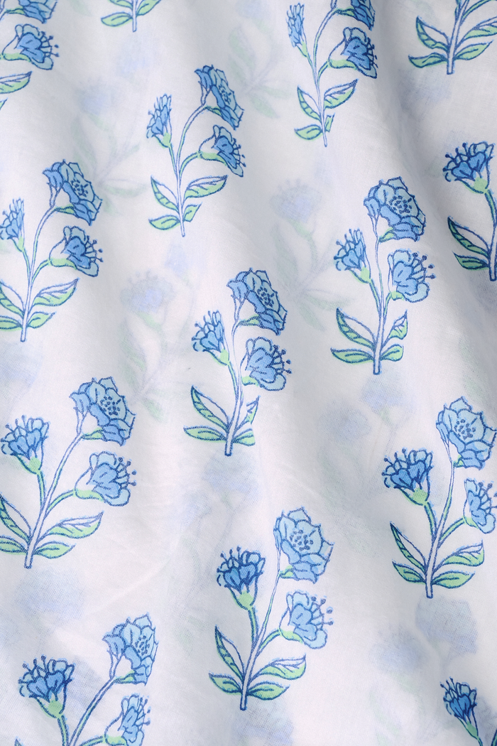 A young girl with long brown hair wears the Roller Rabbit Kids Rosa Ditsy Floral Pami Dress, made from cotton voile with blue-green block print and ruffled sleeves, standing against a plain light background, looking slightly to the side.