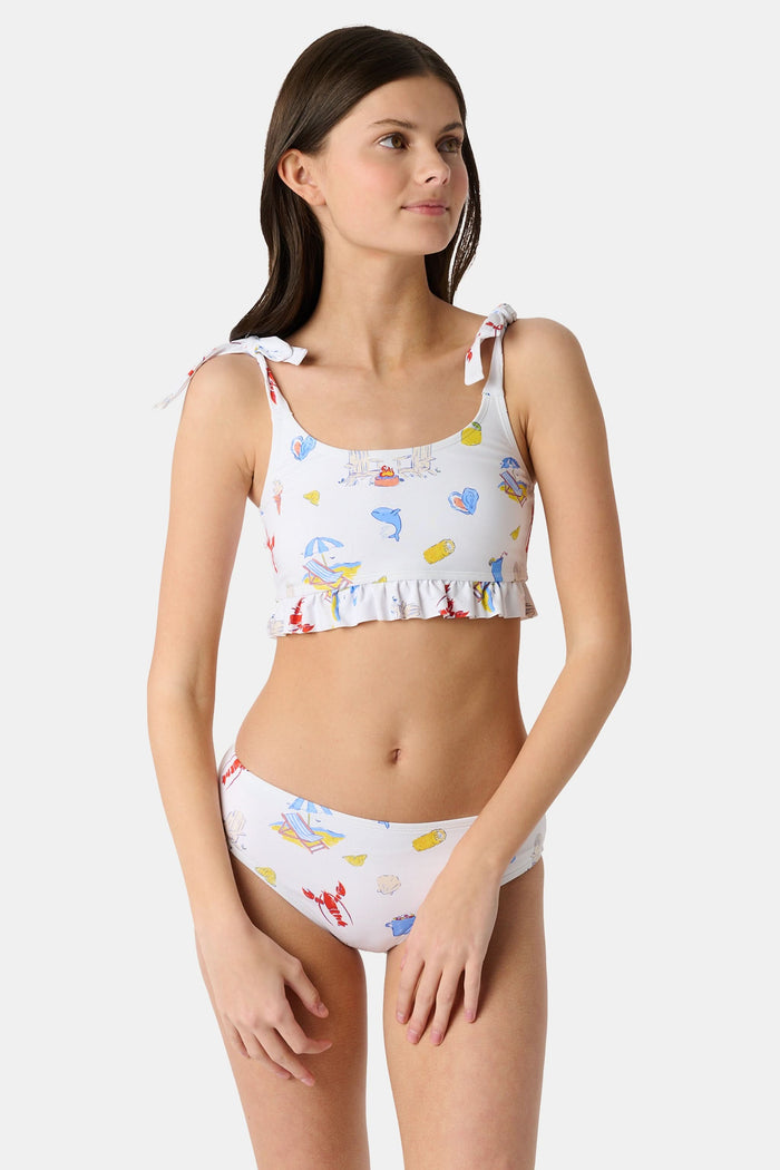 A girl wears the Roller Rabbit Kids Ruffle Catch of the Day Cora Bikini, a white two-piece with a playful print, ruffle trim top, and tie straps, standing against a plain light background.