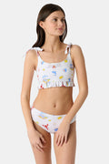 A girl wears the Roller Rabbit Kids Ruffle Catch of the Day Cora Bikini, a white two-piece with a playful print, ruffle trim top, and tie straps, standing against a plain light background.