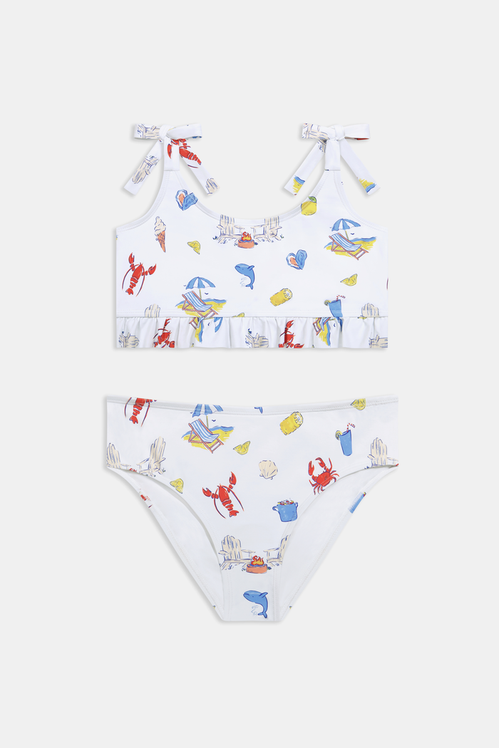 A girl wears the Roller Rabbit Kids Ruffle Catch of the Day Cora Bikini, a white two-piece with a playful print, ruffle trim top, and tie straps, standing against a plain light background.