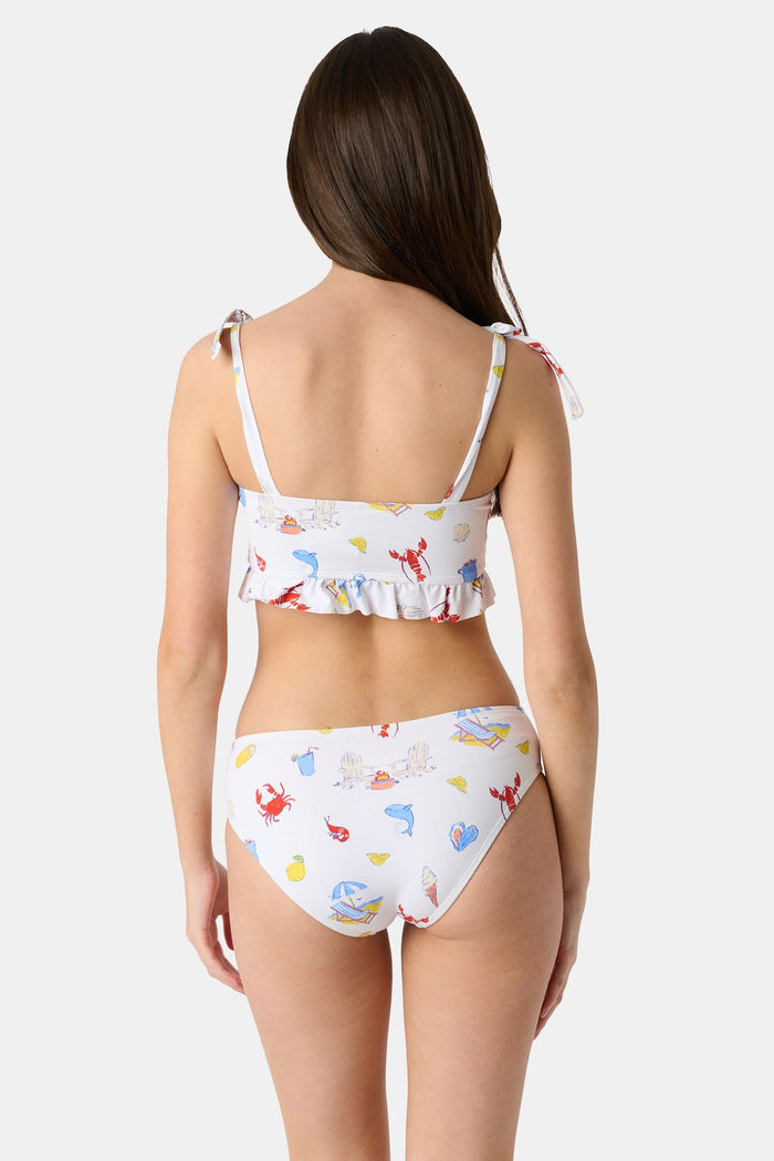 A girl wears the Roller Rabbit Kids Ruffle Catch of the Day Cora Bikini, a white two-piece with a playful print, ruffle trim top, and tie straps, standing against a plain light background.