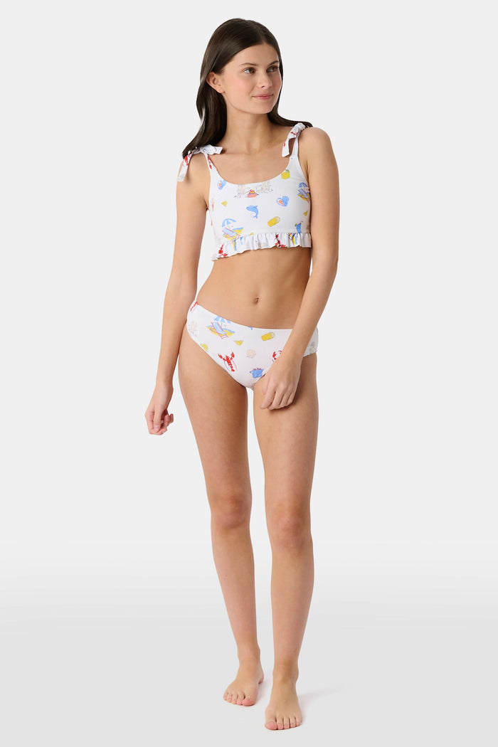 A girl wears the Roller Rabbit Kids Ruffle Catch of the Day Cora Bikini, a white two-piece with a playful print, ruffle trim top, and tie straps, standing against a plain light background.