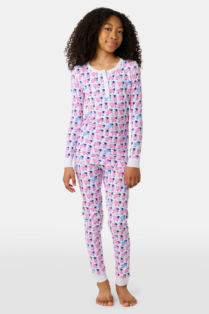 A smiling young girl stands barefoot in Kids Sammy The Sheep Pajamas by Roller Rabbit, posing against a white backdrop to showcase stylish children's sleepwear.
