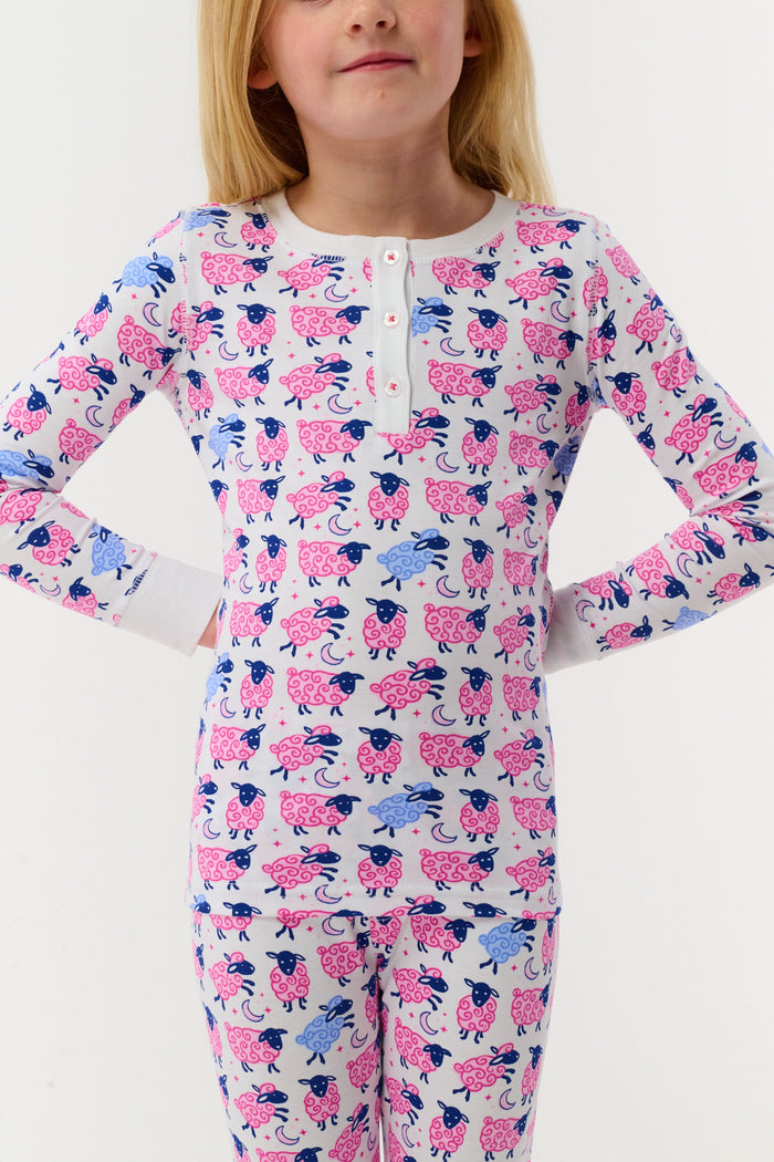 A smiling young girl stands barefoot in Kids Sammy The Sheep Pajamas by Roller Rabbit, posing against a white backdrop to showcase stylish children's sleepwear.
