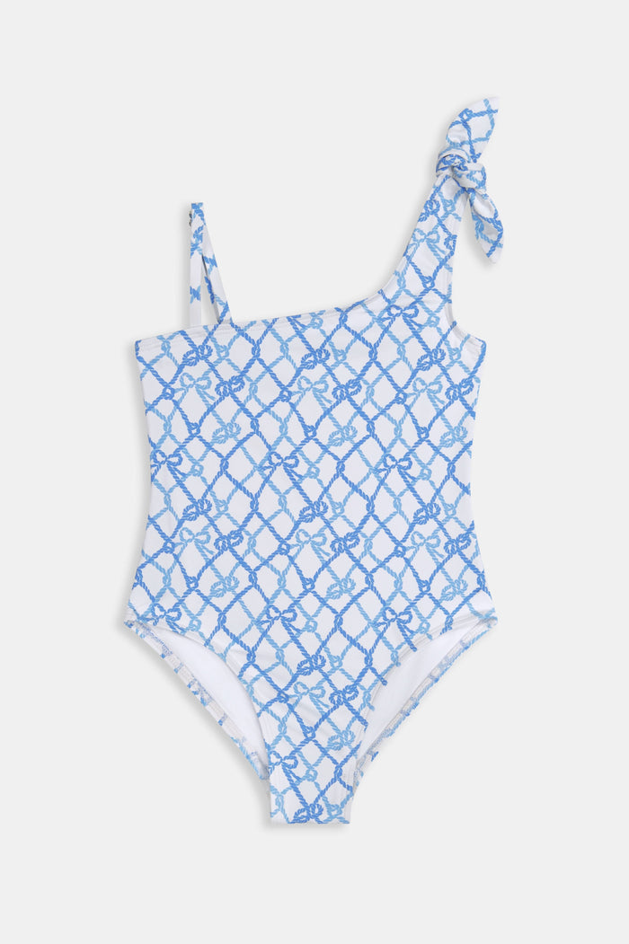 A young girl with long brown hair wears the Roller Rabbit Kids Seaside Knots Jacey One Piece, featuring a blue geometric pattern and a bow on one shoulder, smiling with her hands on her hips against a plain background.