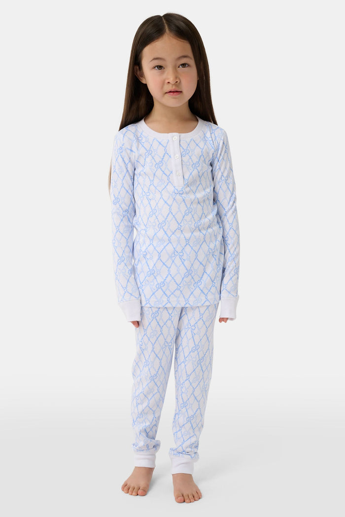 A young girl with long brown hair stands barefoot in Roller Rabbit Kids Seaside Knots Juniper Pajamas, featuring matching white and light blue knot print on soft Pima fabric with long sleeves and button details, against a plain light background.