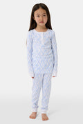 A young girl with long brown hair stands barefoot in Roller Rabbit Kids Seaside Knots Juniper Pajamas, featuring matching white and light blue knot print on soft Pima fabric with long sleeves and button details, against a plain light background.
