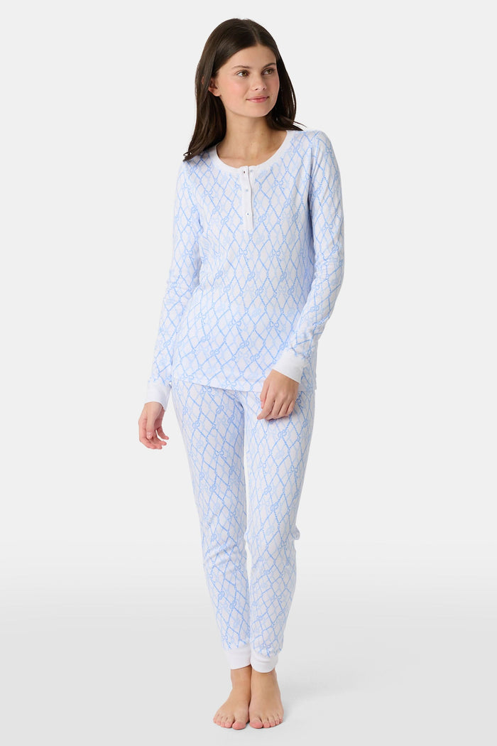 A young girl with long brown hair stands barefoot in Roller Rabbit Kids Seaside Knots Juniper Pajamas, featuring matching white and light blue knot print on soft Pima fabric with long sleeves and button details, against a plain light background.
