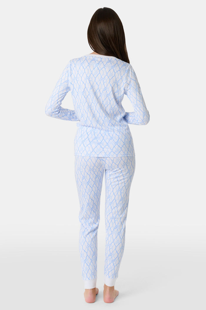 A young girl with long brown hair stands barefoot in Roller Rabbit Kids Seaside Knots Juniper Pajamas, featuring matching white and light blue knot print on soft Pima fabric with long sleeves and button details, against a plain light background.