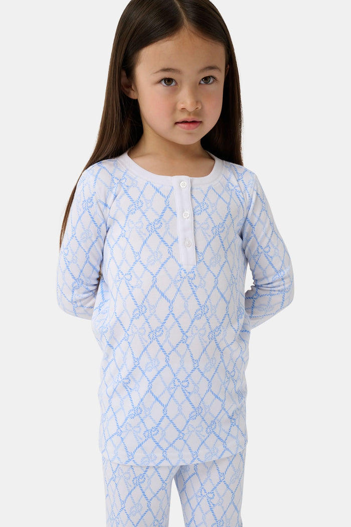 A young girl with long brown hair stands barefoot in Roller Rabbit Kids Seaside Knots Juniper Pajamas, featuring matching white and light blue knot print on soft Pima fabric with long sleeves and button details, against a plain light background.