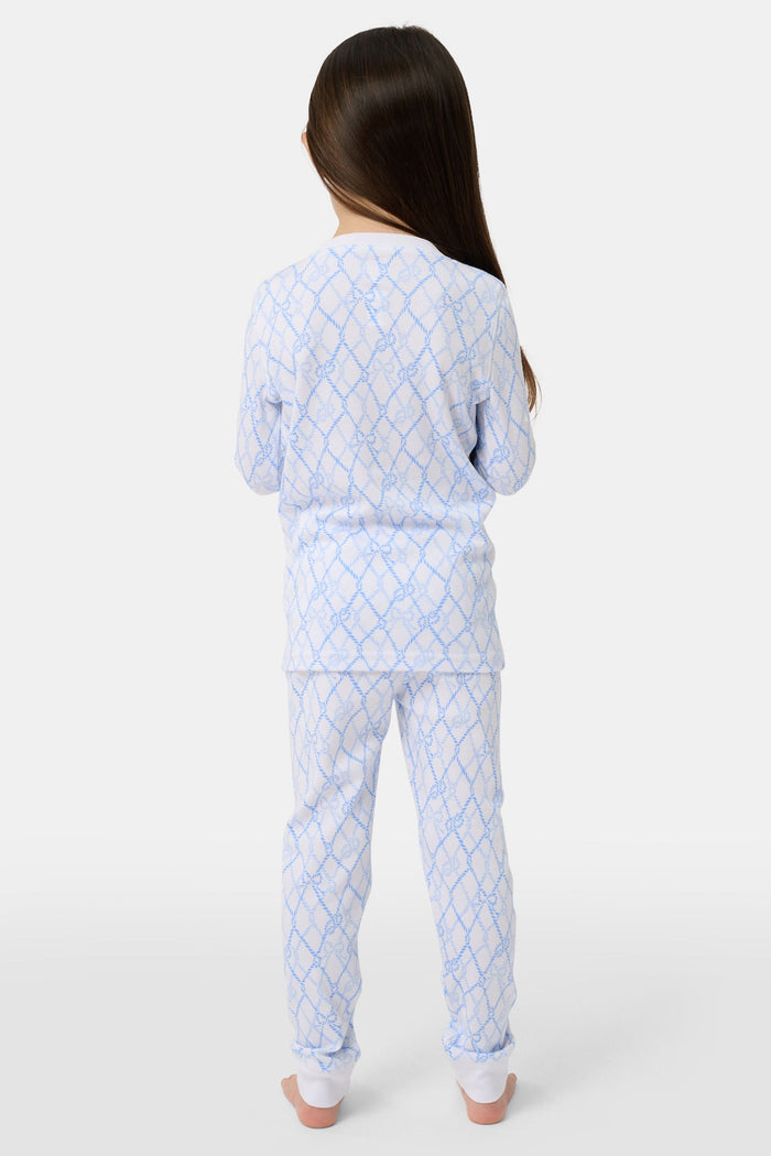 A young girl with long brown hair stands barefoot in Roller Rabbit Kids Seaside Knots Juniper Pajamas, featuring matching white and light blue knot print on soft Pima fabric with long sleeves and button details, against a plain light background.