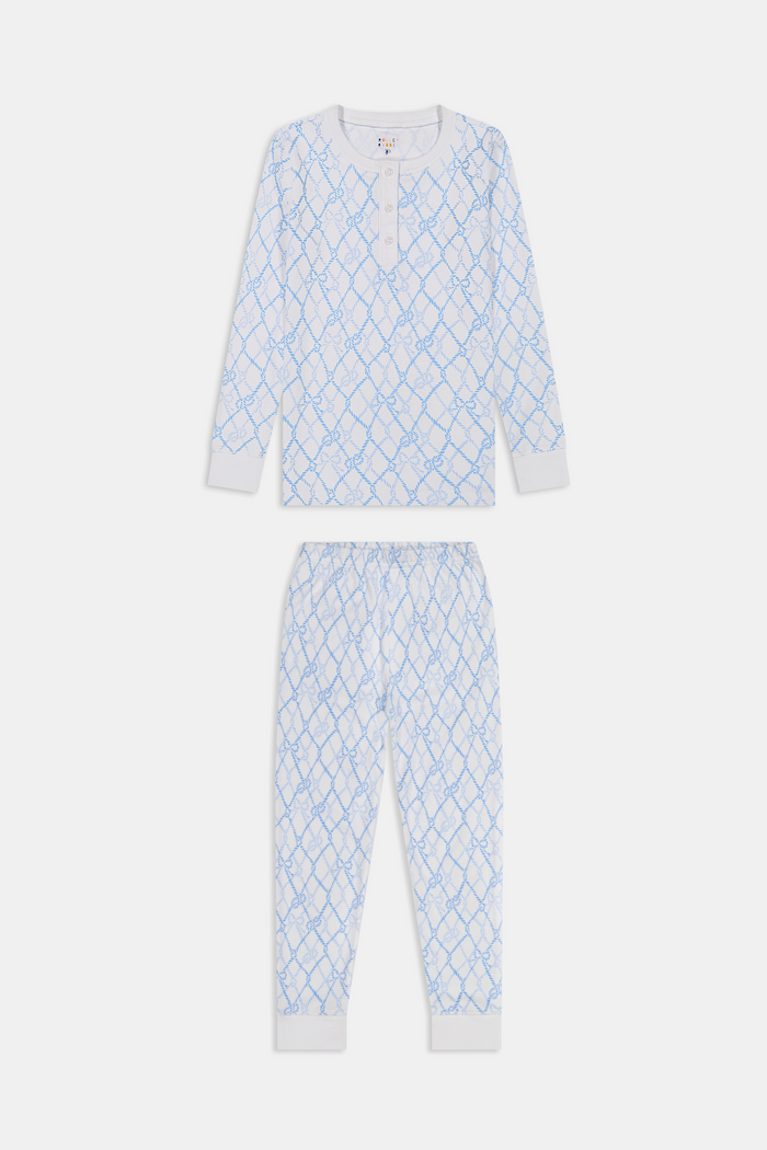 A young girl with long brown hair stands barefoot in Roller Rabbit Kids Seaside Knots Juniper Pajamas, featuring matching white and light blue knot print on soft Pima fabric with long sleeves and button details, against a plain light background.