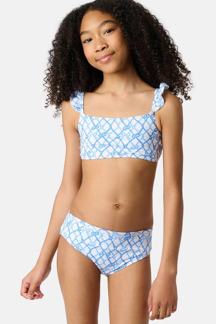 A young girl with curly hair smiles in the Roller Rabbit Kids Seaside Knots Rory Bikini, featuring ruffled shoulder straps and UPF 50+ sun protection, posing against a plain light background.