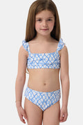 A young girl with curly hair smiles in the Roller Rabbit Kids Seaside Knots Rory Bikini, featuring ruffled shoulder straps and UPF 50+ sun protection, posing against a plain light background.
