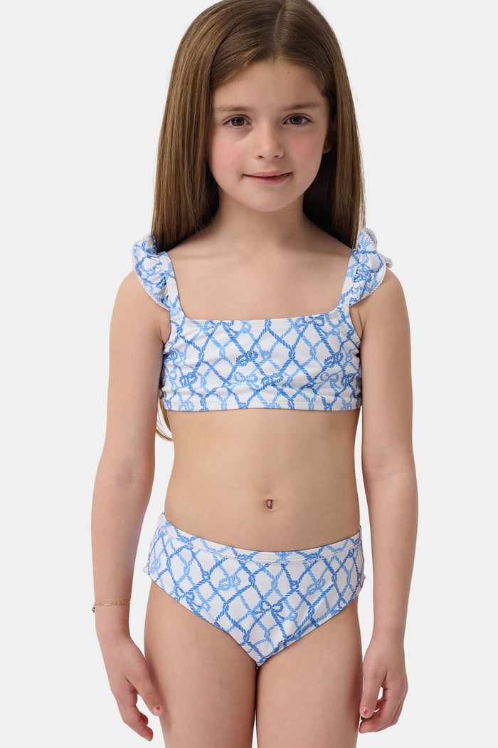 A young girl with curly hair smiles in the Roller Rabbit Kids Seaside Knots Rory Bikini, featuring ruffled shoulder straps and UPF 50+ sun protection, posing against a plain light background.