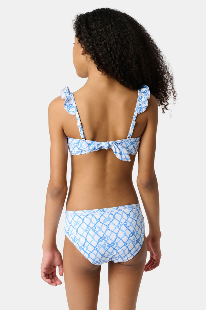 A young girl with curly hair smiles in the Roller Rabbit Kids Seaside Knots Rory Bikini, featuring ruffled shoulder straps and UPF 50+ sun protection, posing against a plain light background.