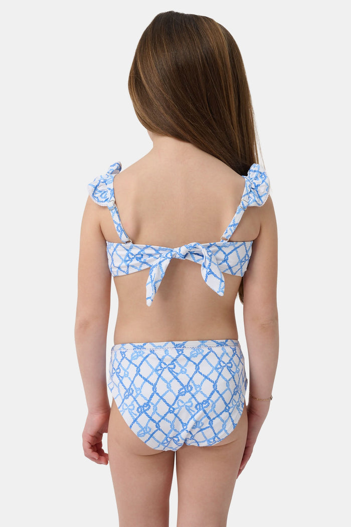A young girl with curly hair smiles in the Roller Rabbit Kids Seaside Knots Rory Bikini, featuring ruffled shoulder straps and UPF 50+ sun protection, posing against a plain light background.