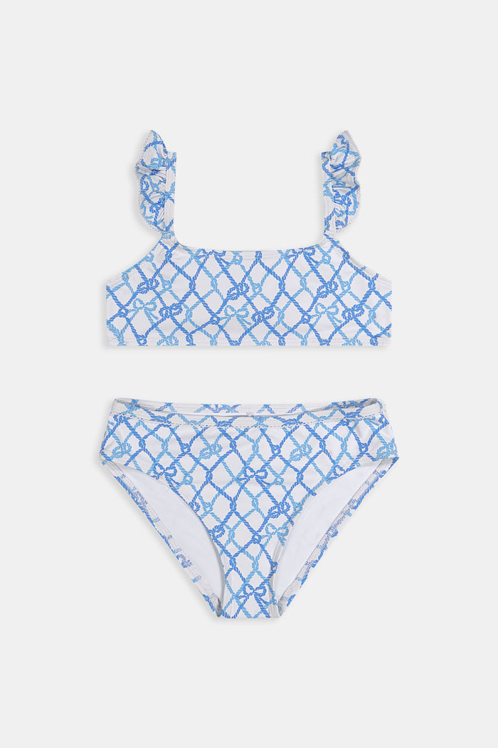 A young girl with curly hair smiles in the Roller Rabbit Kids Seaside Knots Rory Bikini, featuring ruffled shoulder straps and UPF 50+ sun protection, posing against a plain light background.
