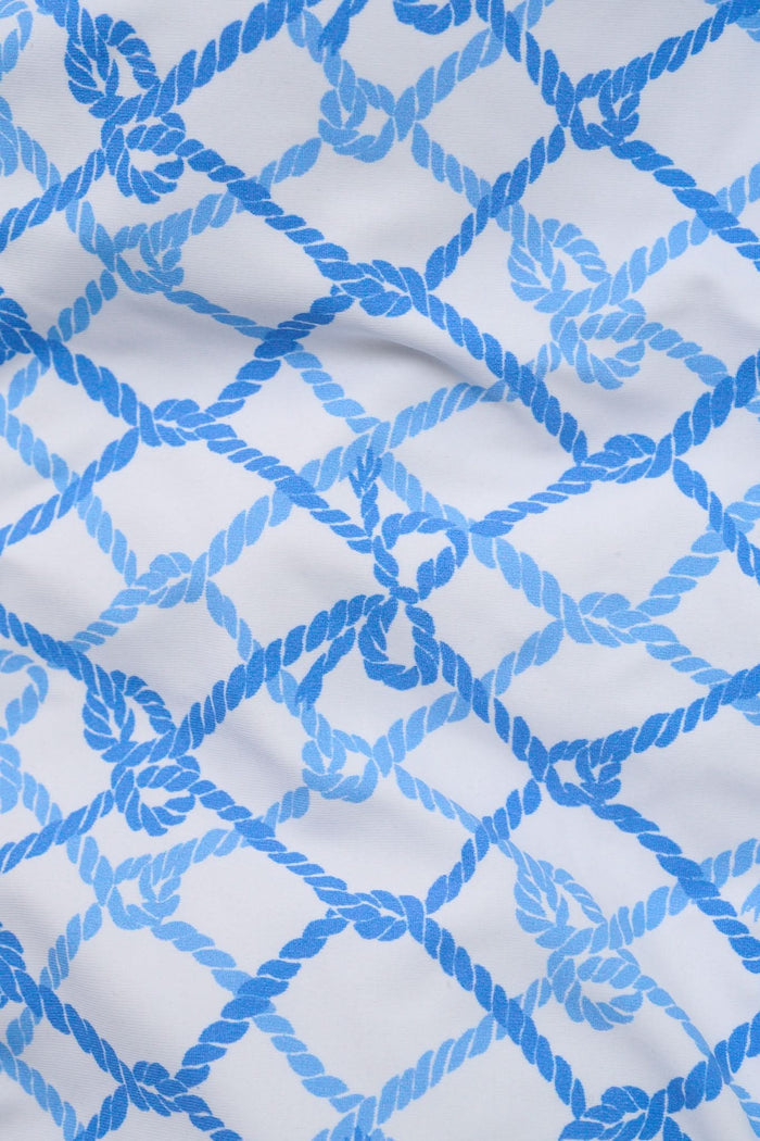 A young girl with long brown hair wears the Roller Rabbit Kids Seaside Knots Jacey One Piece, featuring a blue geometric pattern and a bow on one shoulder, smiling with her hands on her hips against a plain background.